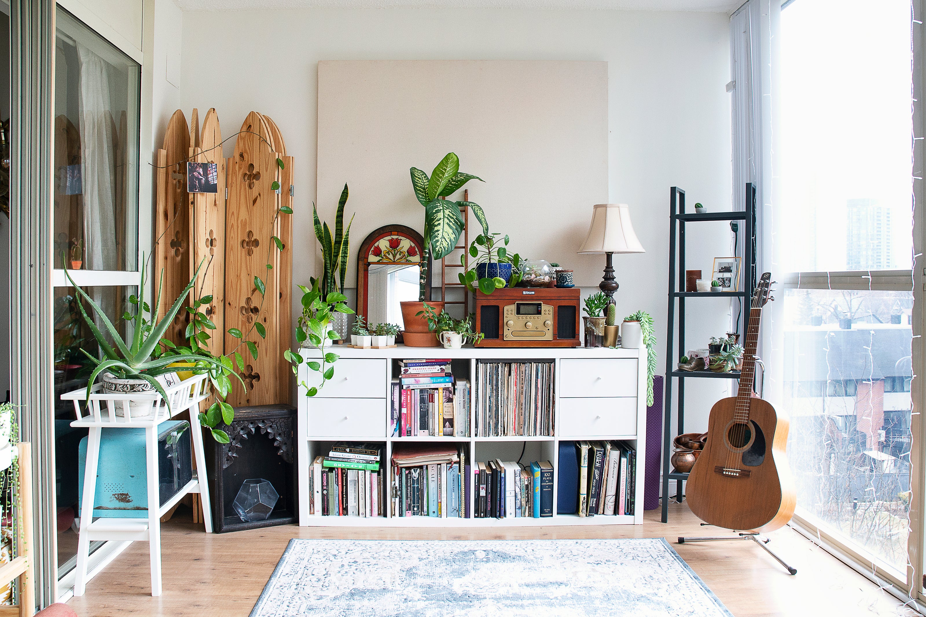 Home Decor with mini showcase having mini flower pot, figurines A guitar in the room. Bookshelf containing books, a lamp on top of it. Plant stand with a potted plant, rug on the floorboard